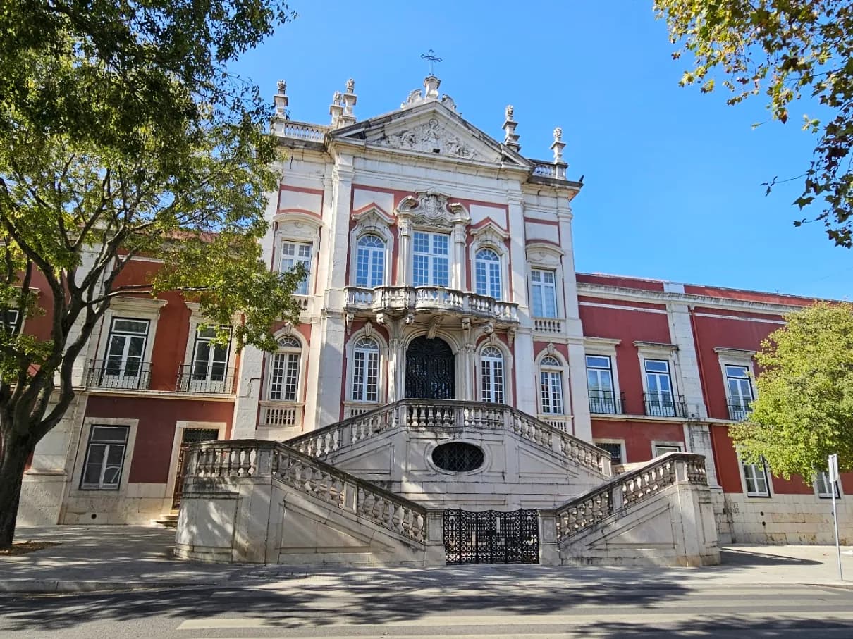 A baroque palace façade in pink-red and white stone under a blue sky, with a double flight of balustraded steps leading up to a central doorway, a balcony above it and a carved pediment topped by a cross. Trees frame the building on both sides.
