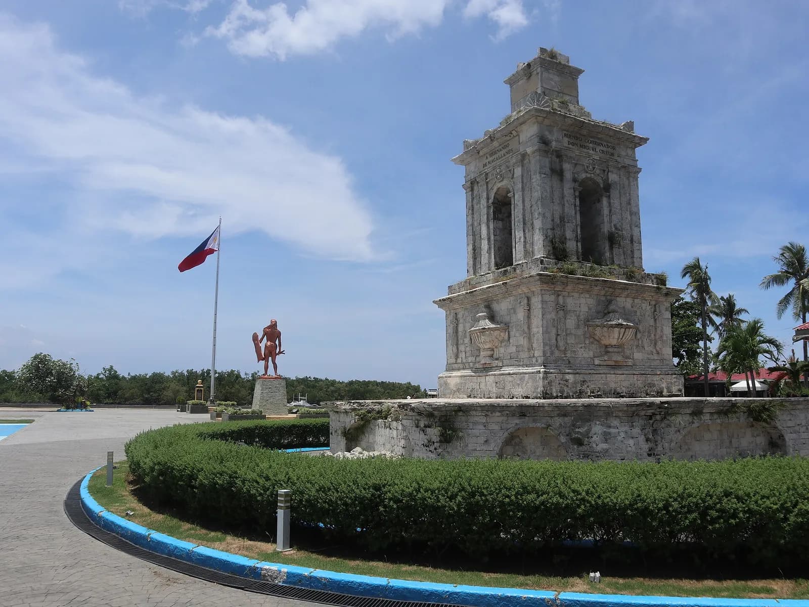 A weathered square stone monument with arched openings and carved urns stands on a low walled base behind a curved hedge. Further back, across a paved plaza, a red-brown statue of a warrior holding a shield and a blade stands on a pedestal near a tall flagpole flying the Philippine flag, under a blue sky.