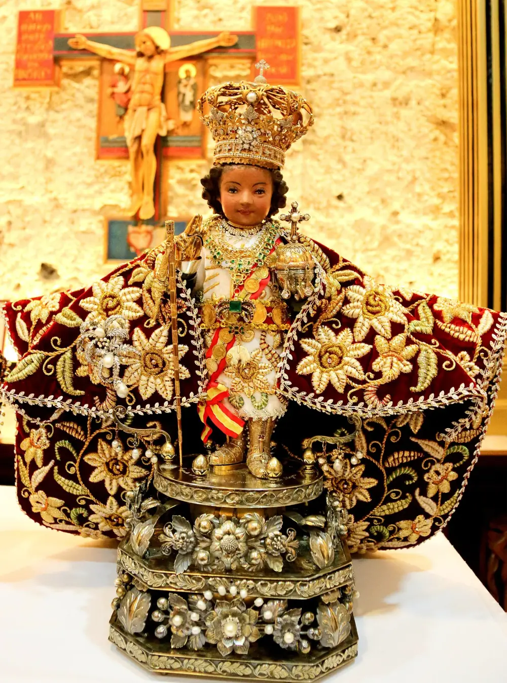 A small dark wooden figure of the child Jesus wearing a jewelled gold crown and a long red cape embroidered with gold flowers stands on an ornate silver base, in front of a wooden crucifix on a stone wall.