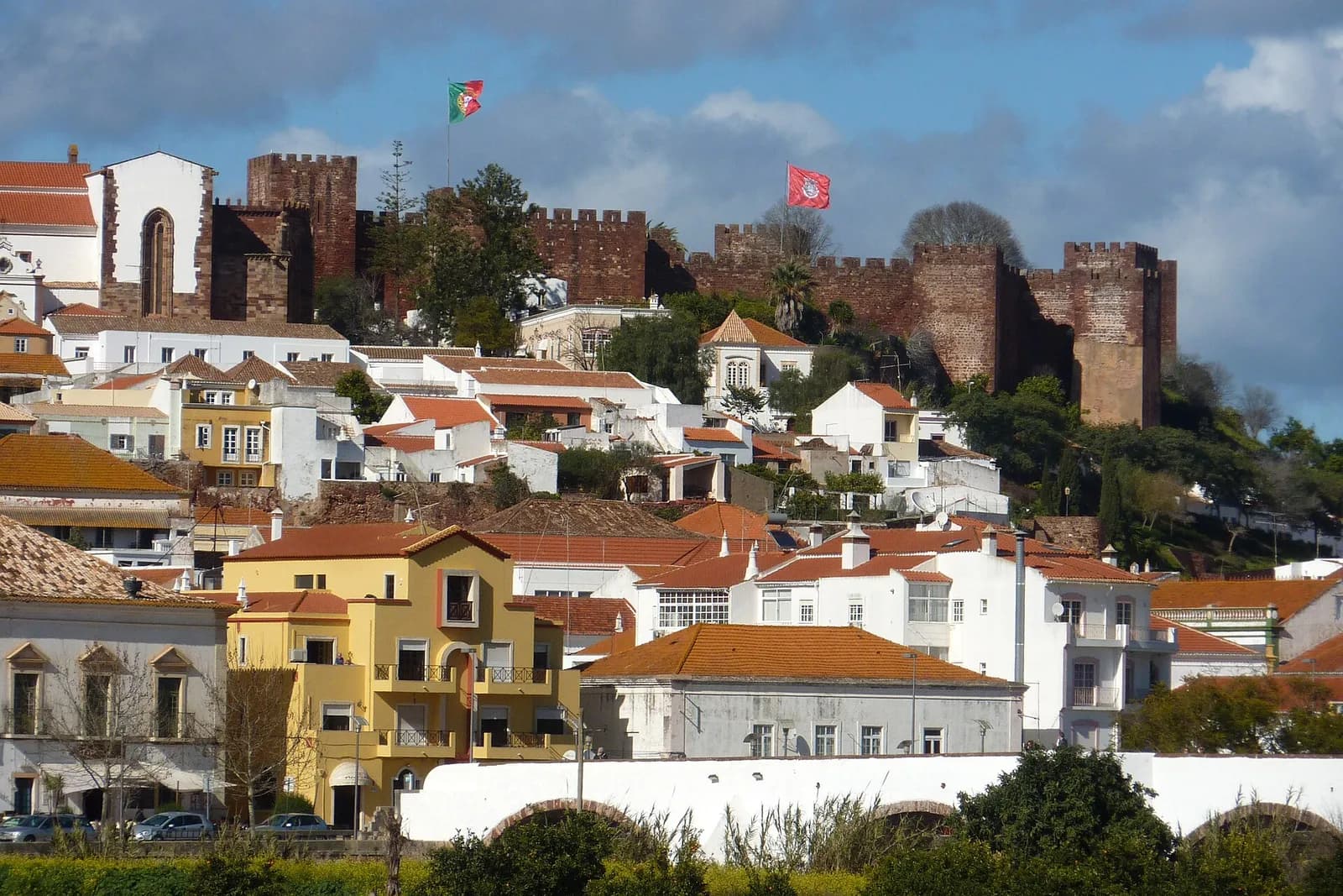 Red sandstone castle walls with square crenellated towers crown a hill above white and yellow houses with orange tiled roofs, under a cloudy blue sky. A Portuguese flag flies on the left and a red flag on the right.