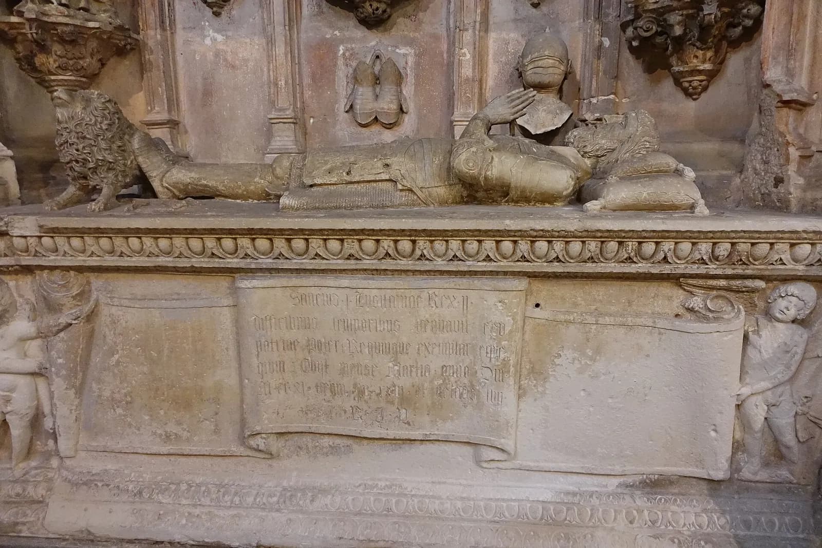 A stone effigy of a king in armour lies on a carved tomb chest, his head on cushions, one hand raised, a lion at his feet and a helmet carved on the wall behind. A Latin inscription in Gothic letters fills a stone scroll on the front of the chest, flanked by carved boys.