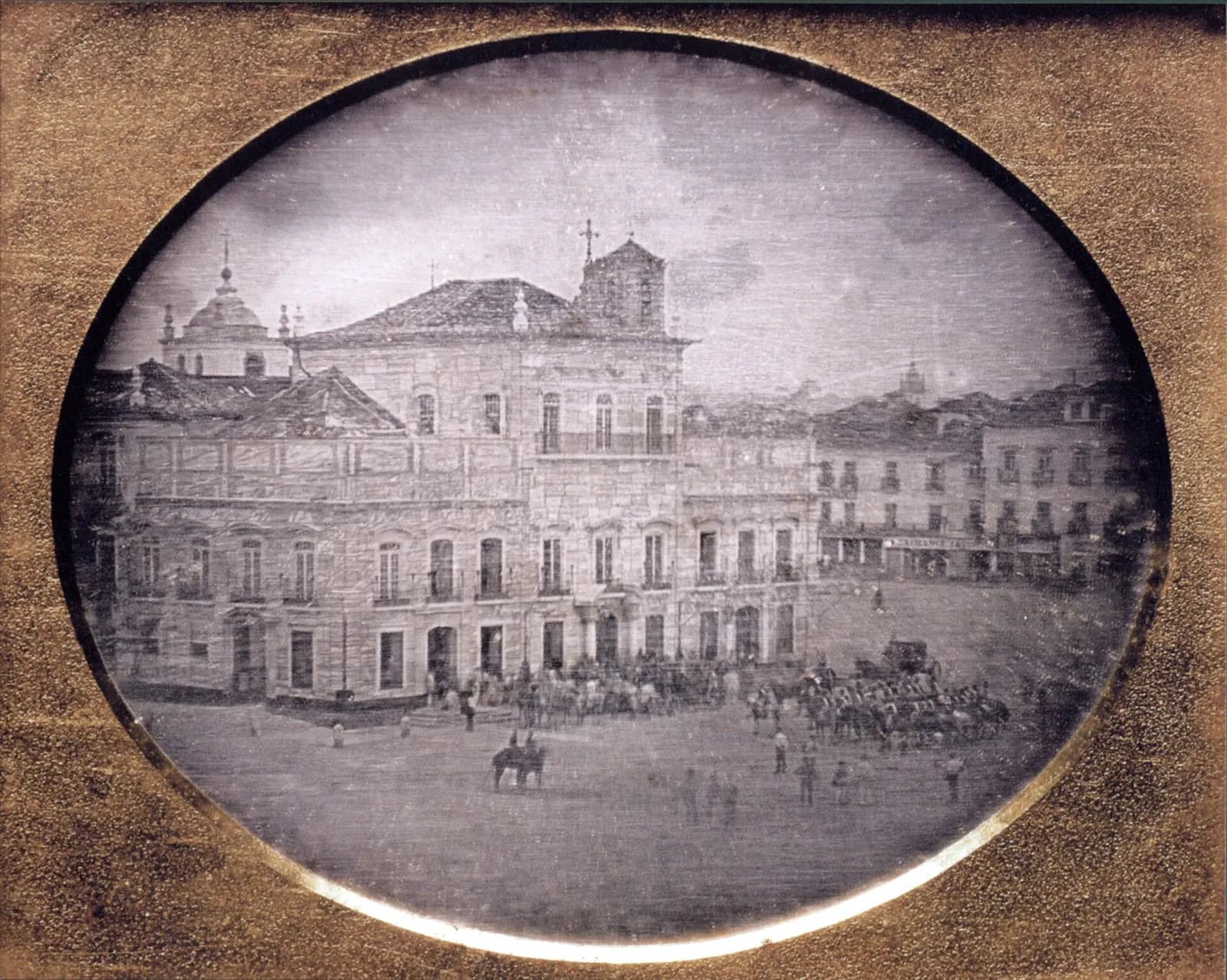 An oval, silvery early photograph in a gilt mount shows a long stone palace with tiled roofs and iron balconies, with a church dome and a bell tower rising behind it, facing a wide open square where mounted soldiers and a carriage are drawn up near the entrance.