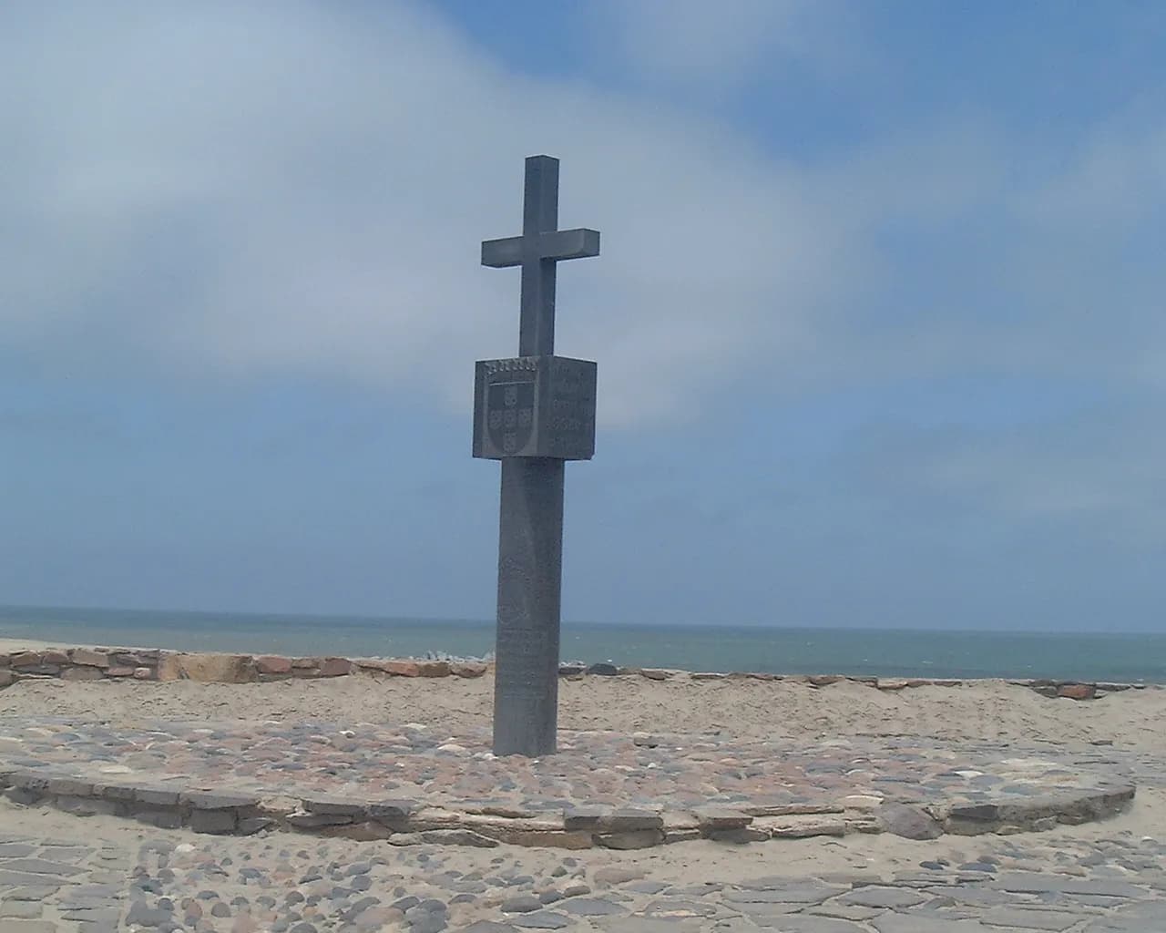 A tall dark stone pillar topped by a cross, with a carved block bearing a coat of arms near the top, stands on a paved circle on a sandy shore. The grey Atlantic and a cloudy sky are behind it.