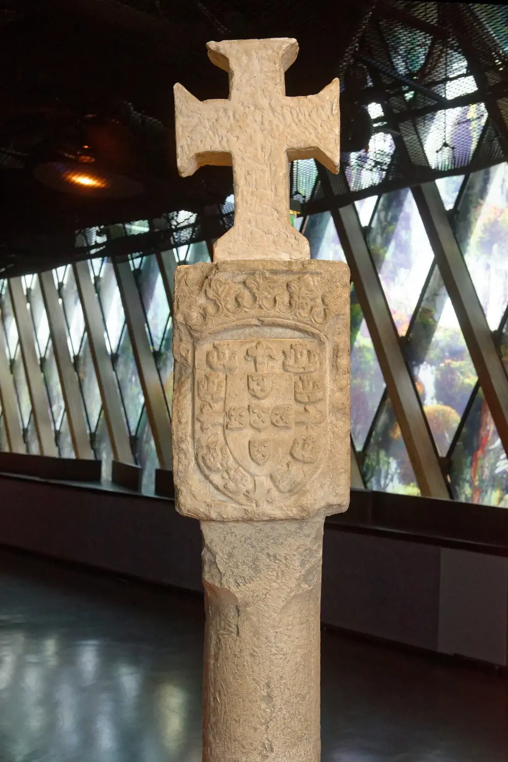 A pale stone-coloured pillar with a square block on top carved with a shield of the Portuguese royal arms, crowned by a stone cross, standing in a dark gallery.