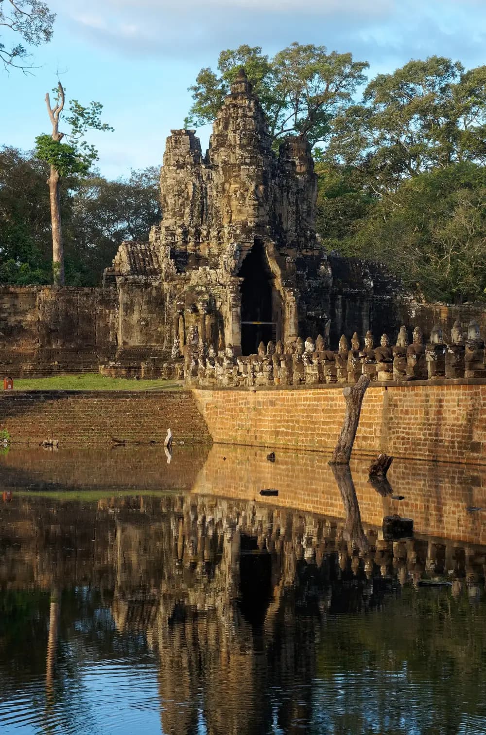 A stone gate tower carved with huge serene faces rises among tall trees. In front, a causeway lined with seated stone figures crosses a moat, and the gate and statues are reflected in the still water.