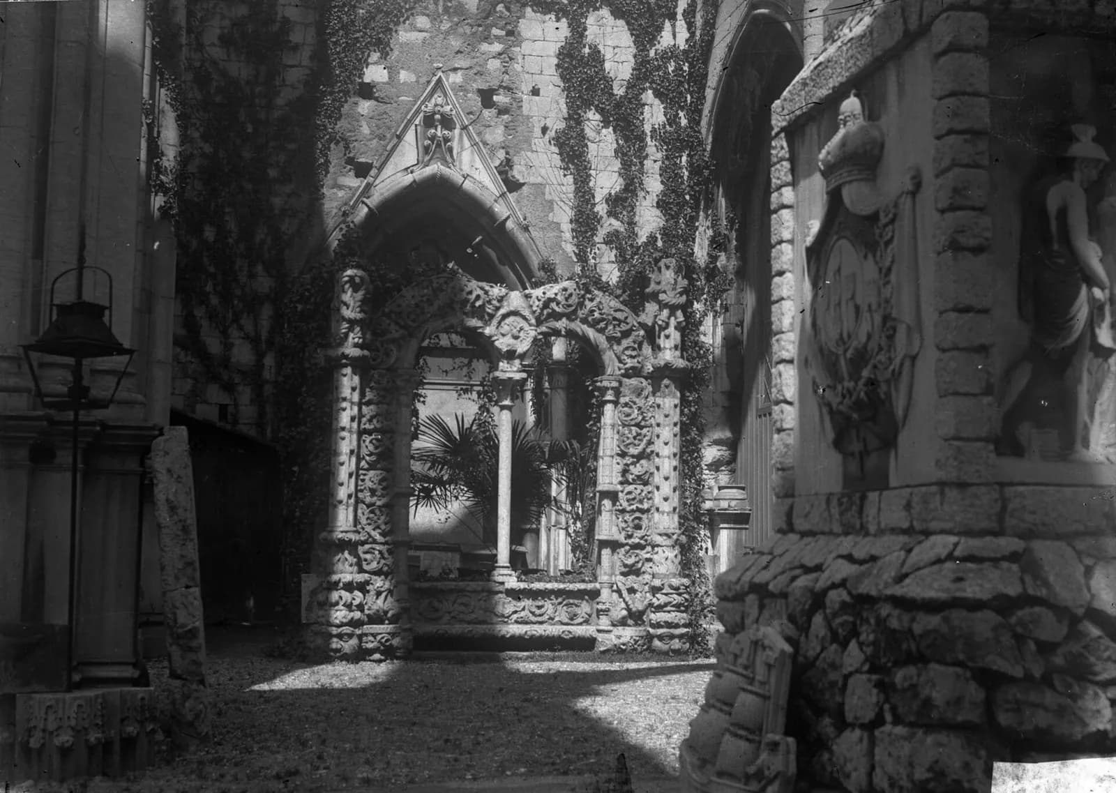 Black-and-white photograph inside a roofless ruin: an ivy-covered Gothic wall, a richly carved stone arcade with twisted columns, a palm behind it, an old street lamp on the left and a carved coat of arms on a stone block on the right.