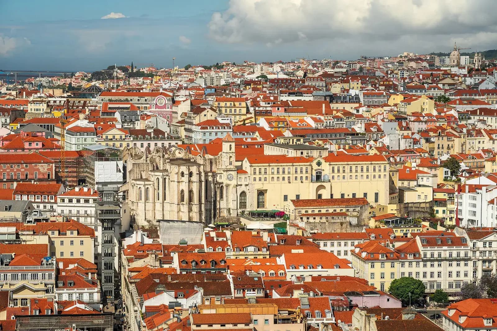 A hillside of red-tiled roofs in central Lisbon. In the middle rise the pale stone walls and buttresses of a roofless Gothic church, with a long yellow building beside it and the iron tower of the Santa Justa lift to the left.