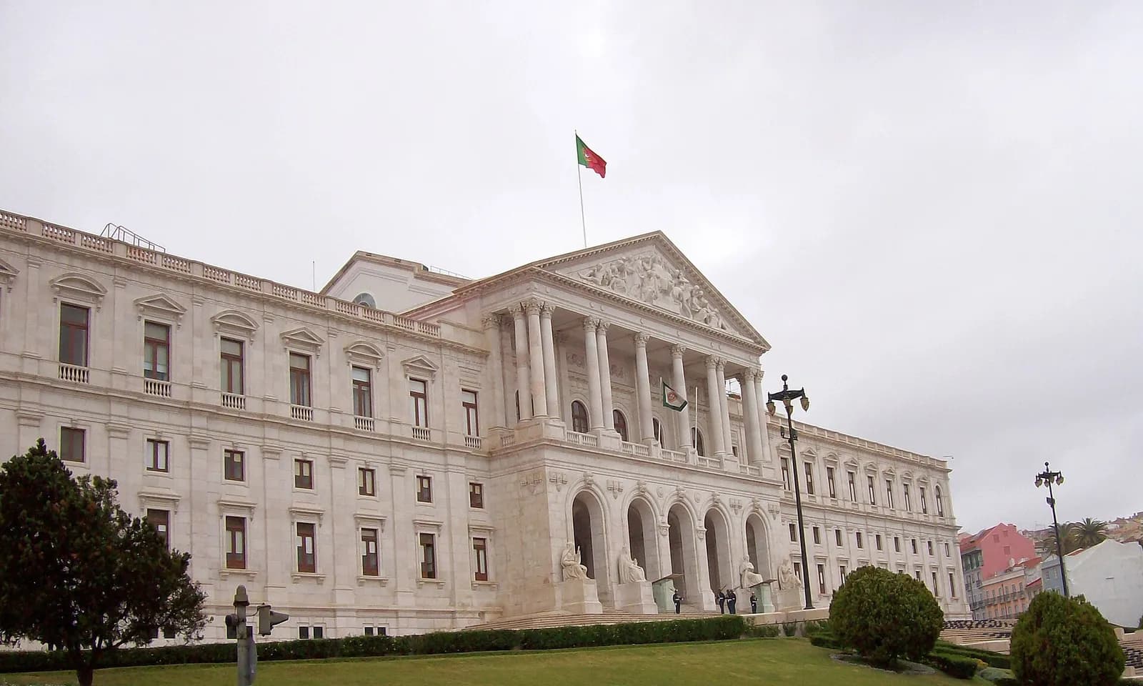 A long white neoclassical palace with a columned portico and pediment, a Portuguese flag flying above it, behind a sloping lawn under a grey sky.