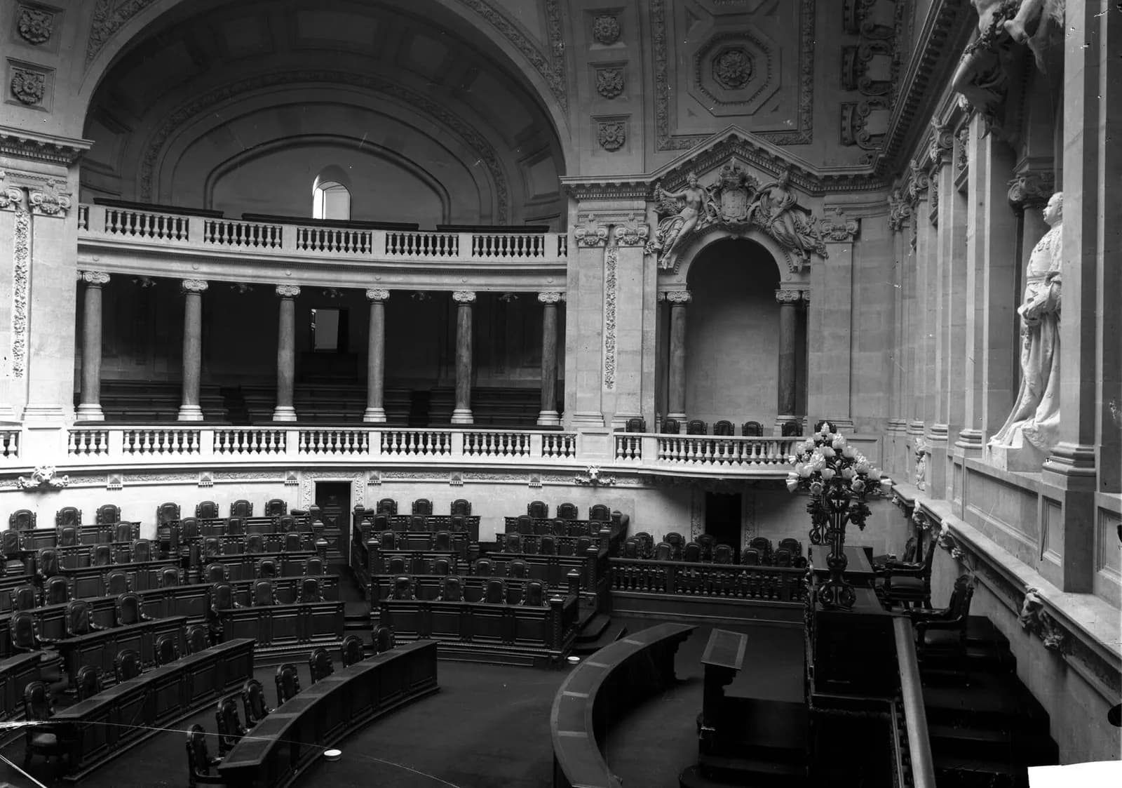 Black-and-white photograph of an empty semicircular debating chamber with rows of wooden desks and leather chairs, a colonnaded gallery above and a marble statue in a niche on the right.
