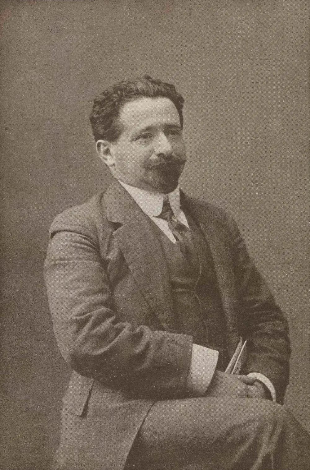 A studio photograph of a seated man with thick dark wavy hair, a full moustache and small beard, wearing a three-piece suit with a high white collar and holding papers on his knee.