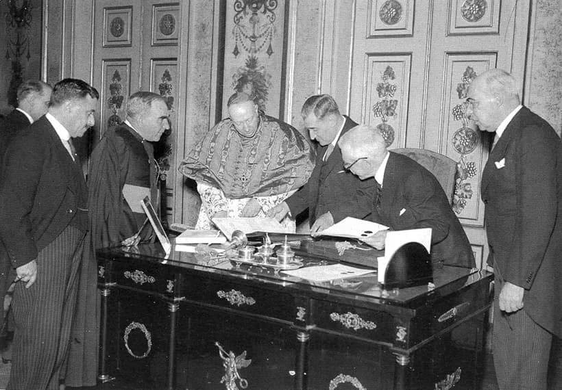 In a room with painted panelled walls, a bald man in glasses sits at an ornate desk reading a document, while a prelate in a patterned cape and several men in dark suits stand around him.
