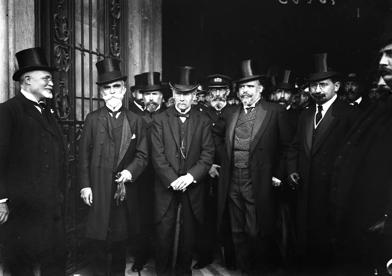 A group of men in dark frock coats and top hats stands in front of a tall wrought-iron gate, with more men crowding behind them. Several have white beards or heavy moustaches; the man in the centre rests both hands on a furled umbrella.