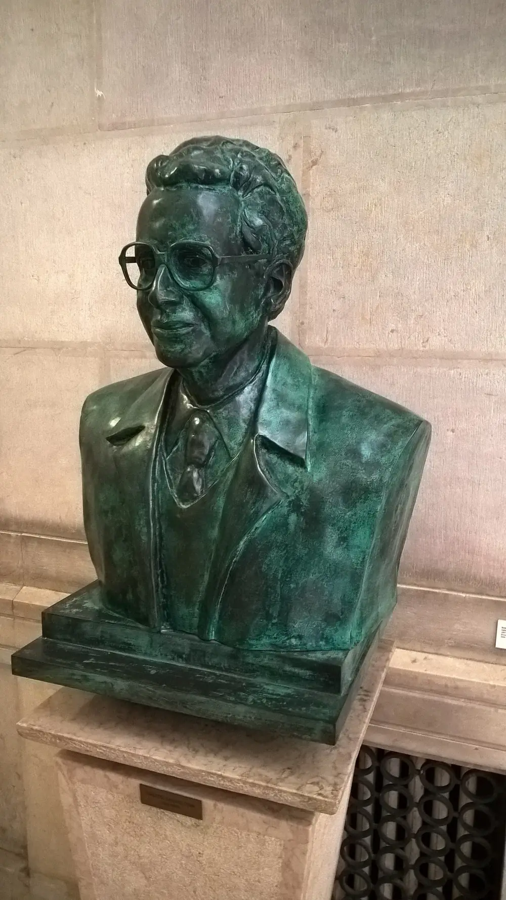A green-patinated bronze bust of a man with wavy hair and large square glasses, wearing a jacket, shirt and tie, on a stone pedestal against a pale stone wall.