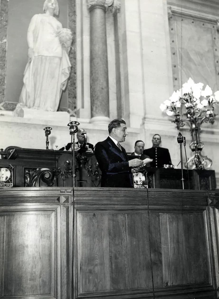Black-and-white photograph of a man in a dark suit speaking from a carved wooden rostrum fitted with several microphones, a sheet of paper in his hand. Officials sit behind him, a vase of white flowers stands to one side and a marble statue fills a niche above.