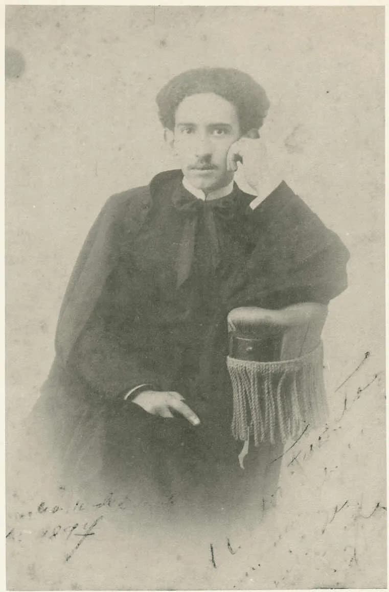Faded studio photograph of a young man with a moustache and dark hair, seated with one arm on a chair with a fringed cushion. He wears a long black cape over a dark cassock closed to the neck, with a white collar and a dark bow at the throat. Handwriting runs across the bottom of the print.