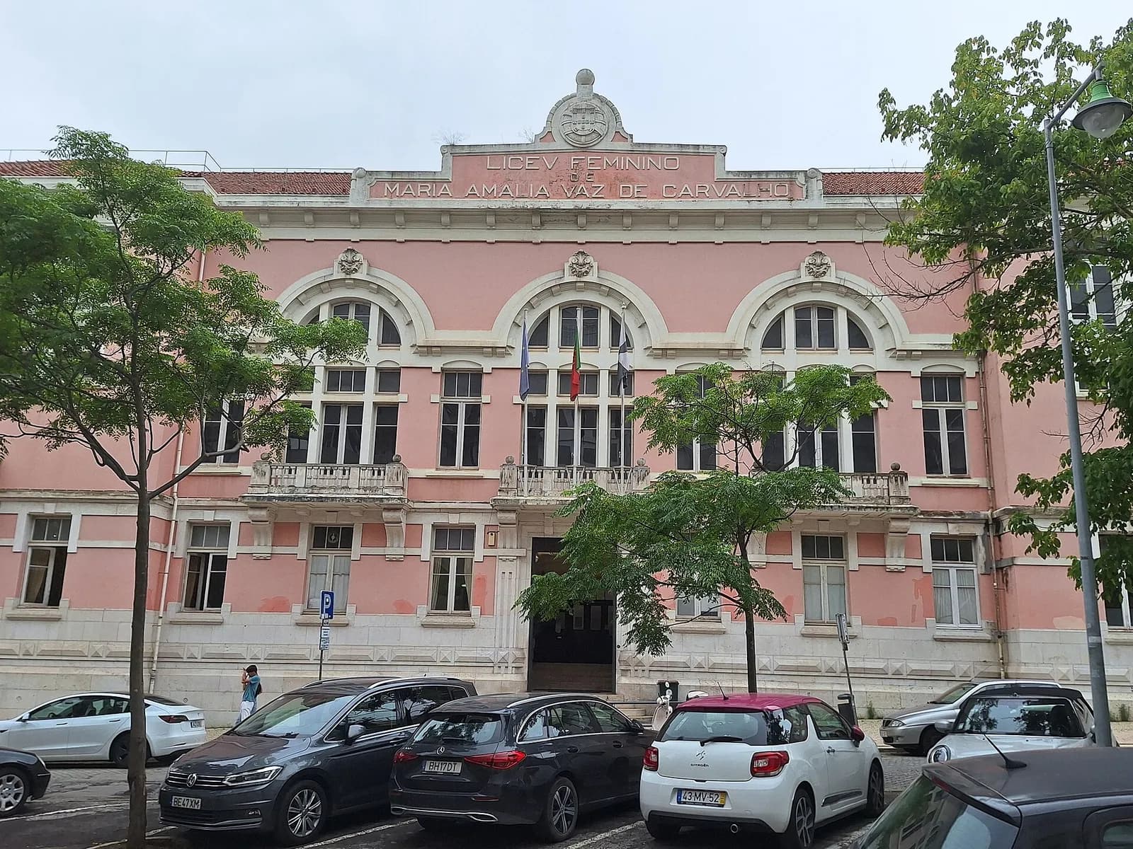 The pink and white front of a two-storey school building with three large arched windows and stone balconies. Lettering at the top reads Liceu Feminino de Maria Amália Vaz de Carvalho beneath a carved coat of arms. Cars are parked in front and trees line the street.
