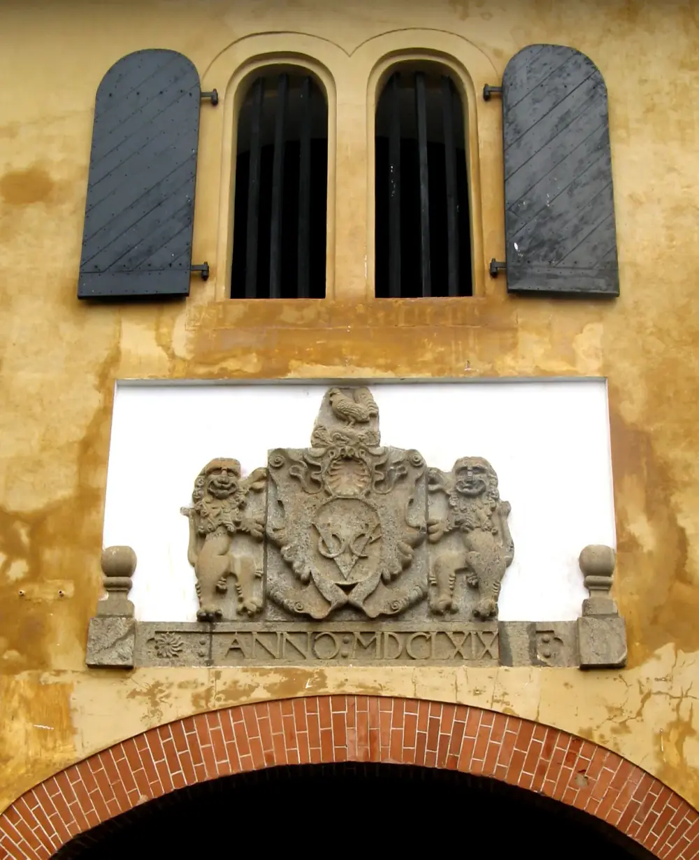 A carved stone coat of arms set into an ochre-washed wall above a brick arch: two lions hold a shield with the interlaced letters VOC, a cockerel perches on top, and a band below reads ANNO MDCLXIX. Two barred arched windows with black shutters are above.