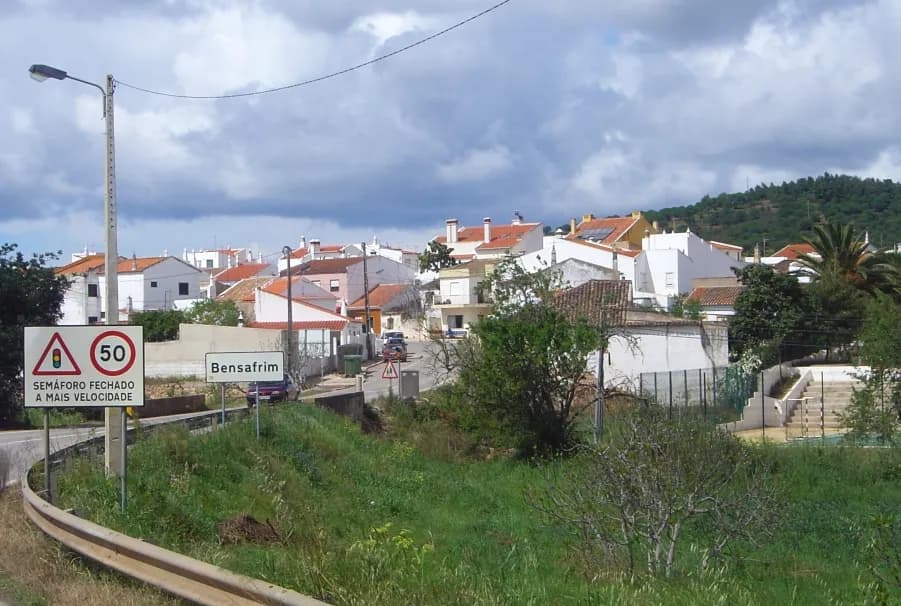 The edge of a small village of white houses with orange-tiled roofs on a slope under a cloudy sky, seen from a road with a sign reading Bensafrim and a speed limit sign.