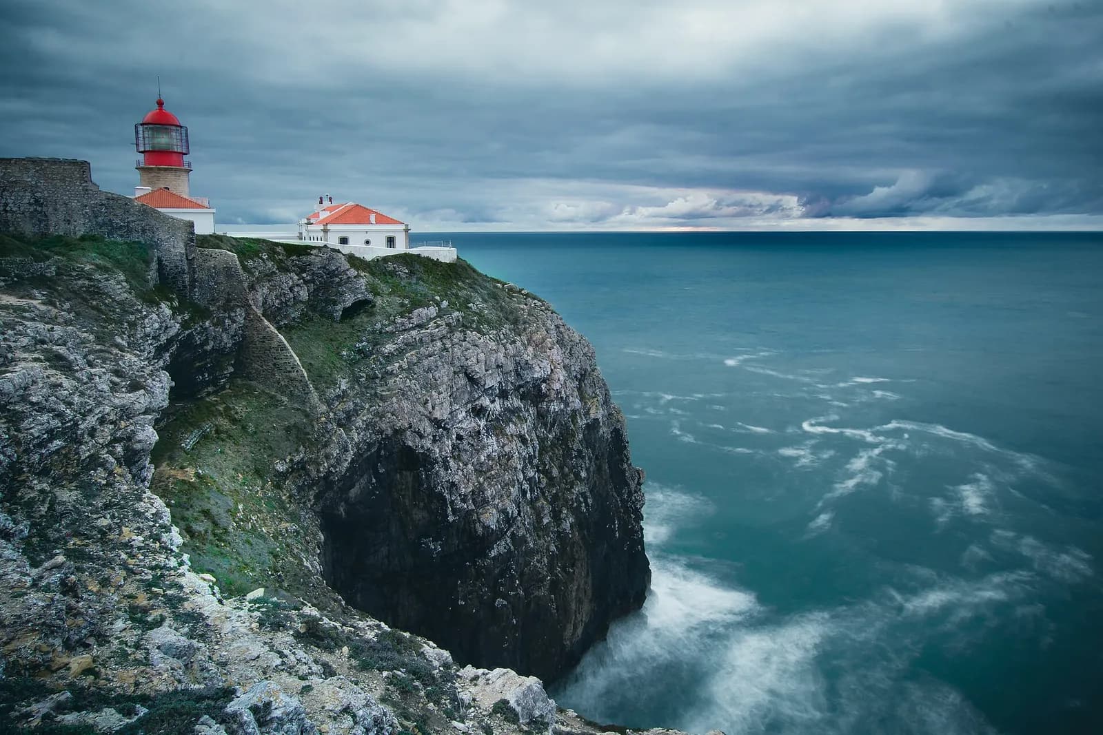 A lighthouse with a red lantern and white buildings with orange roofs stands on top of a sheer grey limestone cliff. Below, the sea swirls white against the rock under a heavy grey sky.