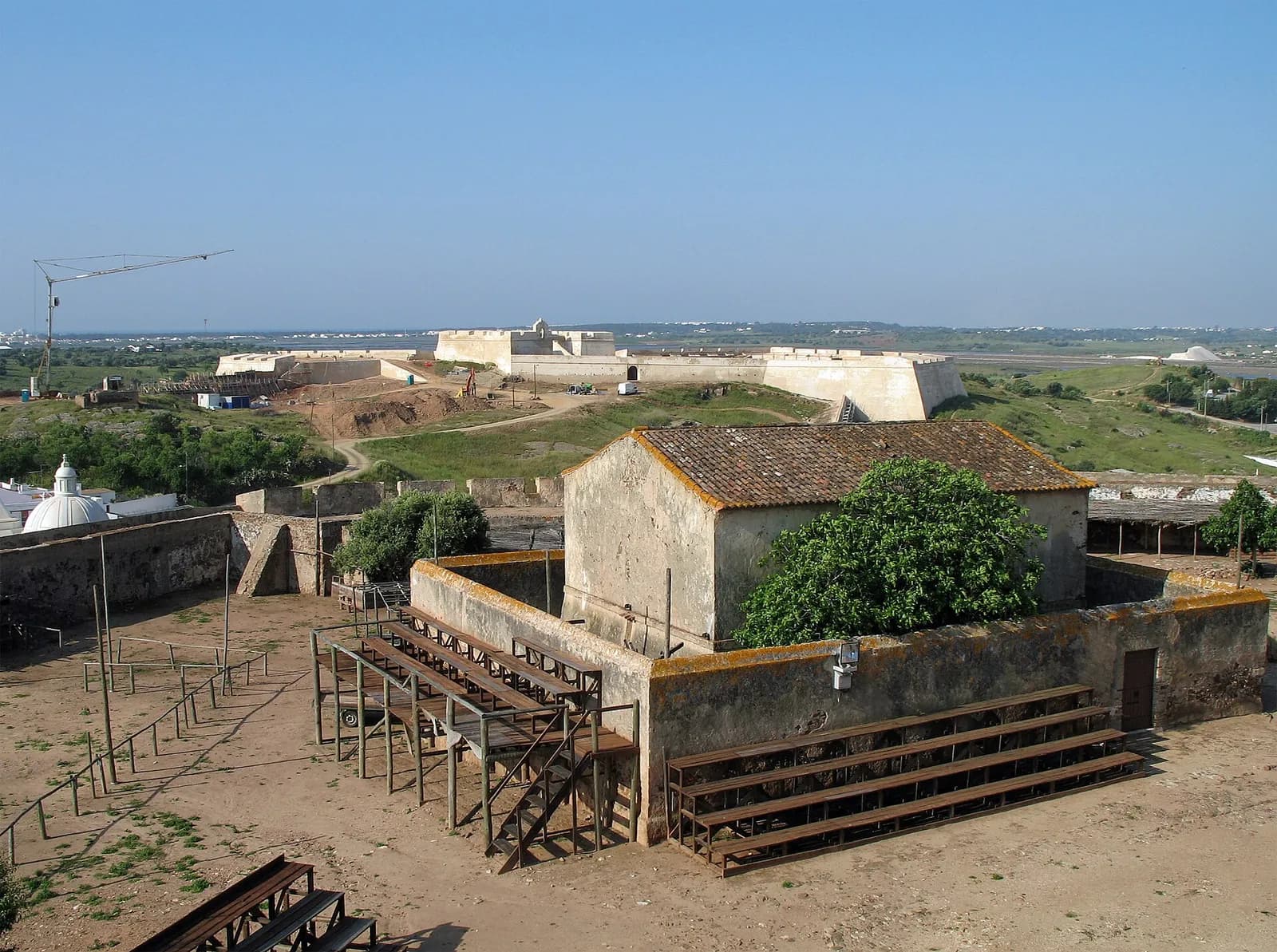 Inside an old castle: a dusty courtyard with wooden benches and an old plastered building with a tiled roof, enclosed by low stone walls. Beyond, on a green hill, stands a low star-shaped fort with white walls, and salt pans and flat country stretch to the horizon.