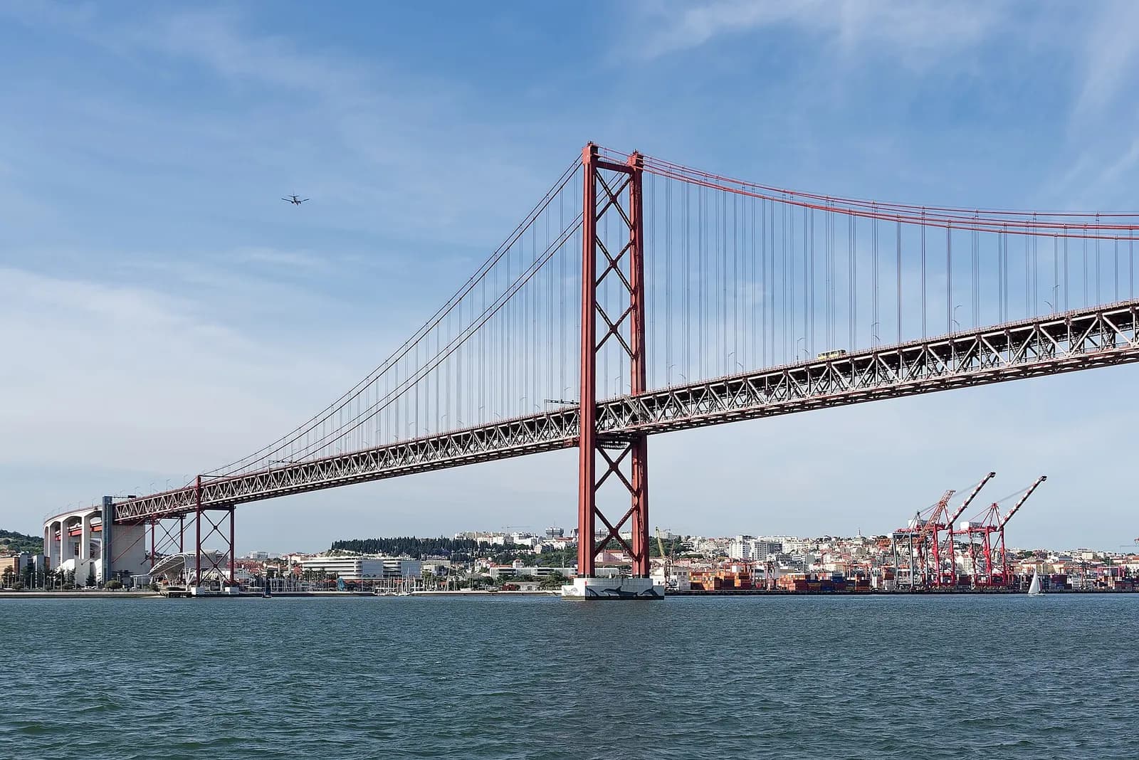 A red suspension bridge seen from the water: a tall lattice tower carries the main cables, which run down to a massive white concrete structure on the shore at left. Behind the bridge are the Lisbon waterfront, port cranes and hillside houses.