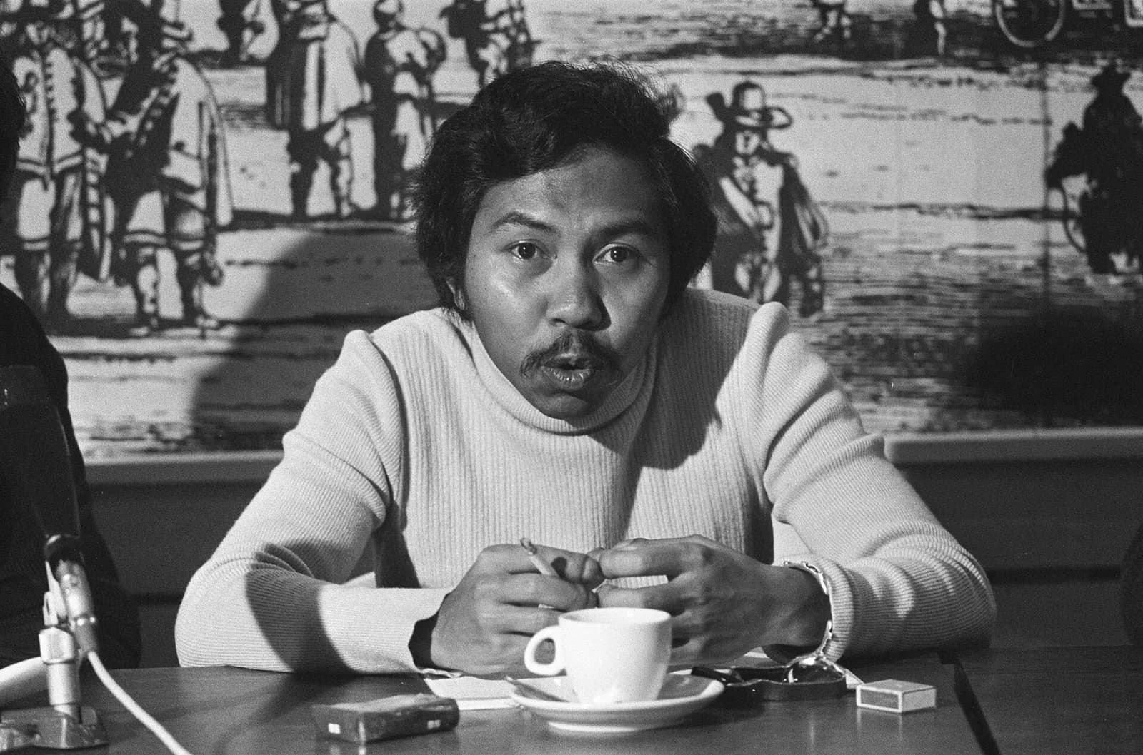 Black-and-white photograph of a young man with a moustache and thick dark hair, wearing a white roll-neck sweater, speaking at a table with a coffee cup and a microphone in front of him and a mural of figures in old-fashioned dress behind.