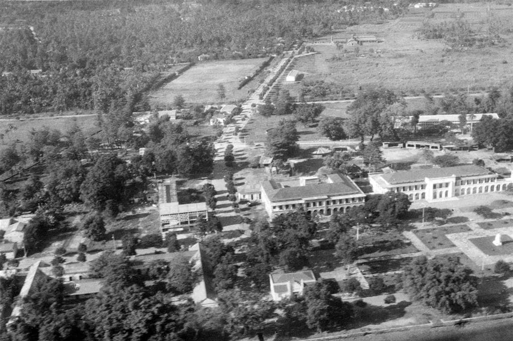 Black-and-white aerial photograph of a small town of low buildings among dense trees, with two large colonial buildings in the centre, an avenue leading inland and open fields and palm groves beyond.