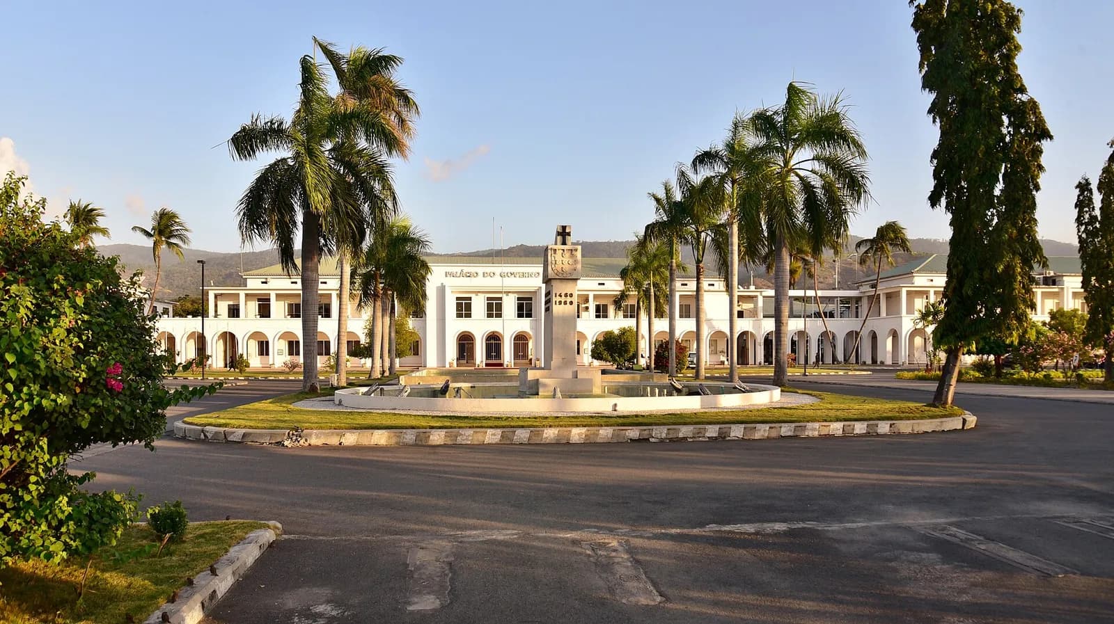 A long white two-storey colonial building with arched arcades and a green roof, its facade lettered Palácio do Governo, seen across a round lawn with a fountain and a stone column, framed by tall palm trees in late afternoon light.