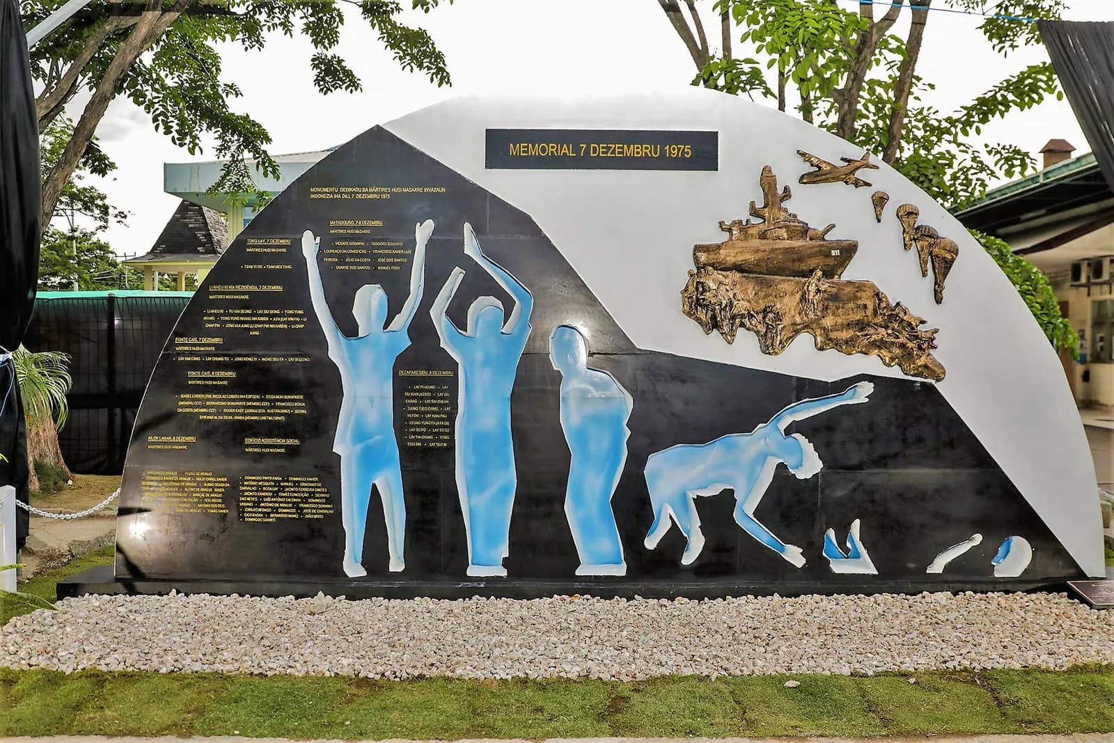 A large half-moon shaped monument in black and white stone. Pale blue figures with raised arms stand and fall across it, beside lists of names in gold lettering, under a bronze relief of a warship, aircraft and parachutes and the heading Memorial 7 Dezembru 1975.