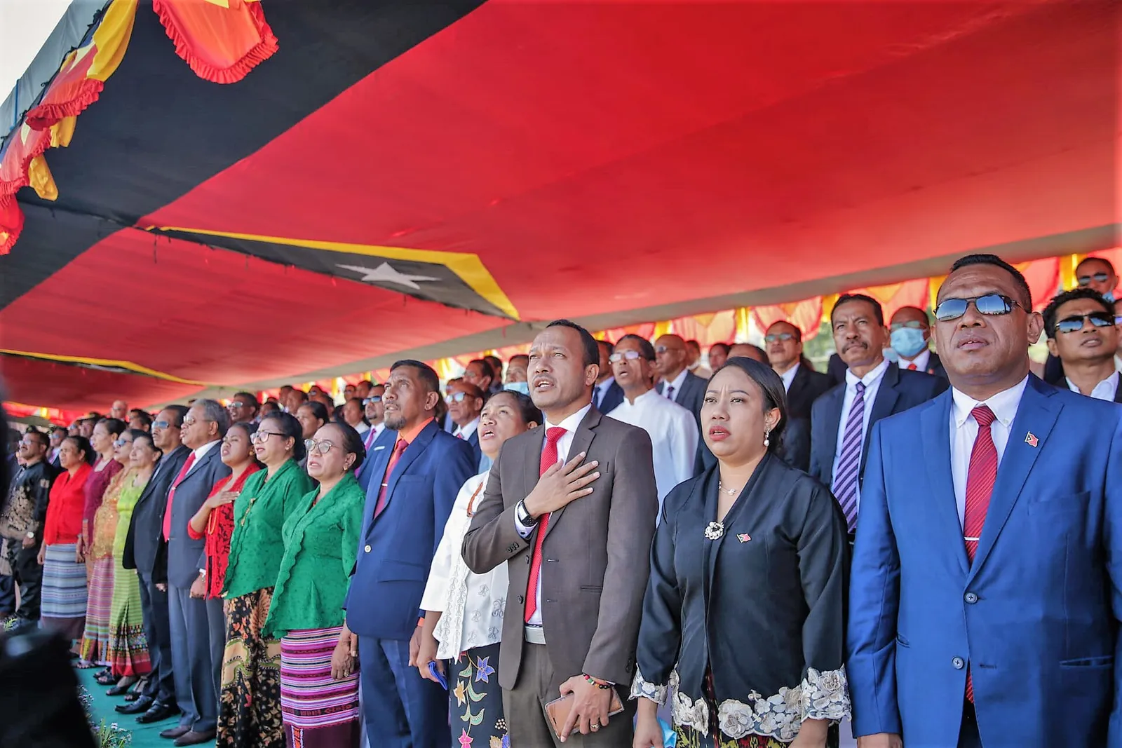 Rows of dignitaries in suits and traditional woven cloth stand under a red awning hung with red, black and yellow bunting, several with a hand on the chest as they sing.