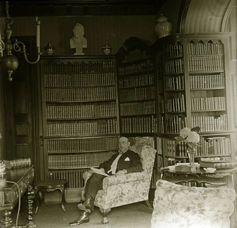 A man in a dark suit with a white pocket handkerchief sits in a flowered armchair with a paper on his lap, in a room lined floor to ceiling with bookcases, a bust on top of one of them and a vase of hydrangeas on a side table.