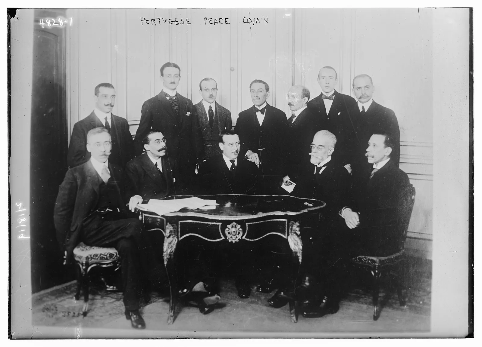 Twelve men in dark suits pose in a panelled room, five seated around an ornate table with papers on it and seven standing behind them. A handwritten label at the top reads Portugese Peace Com'n.