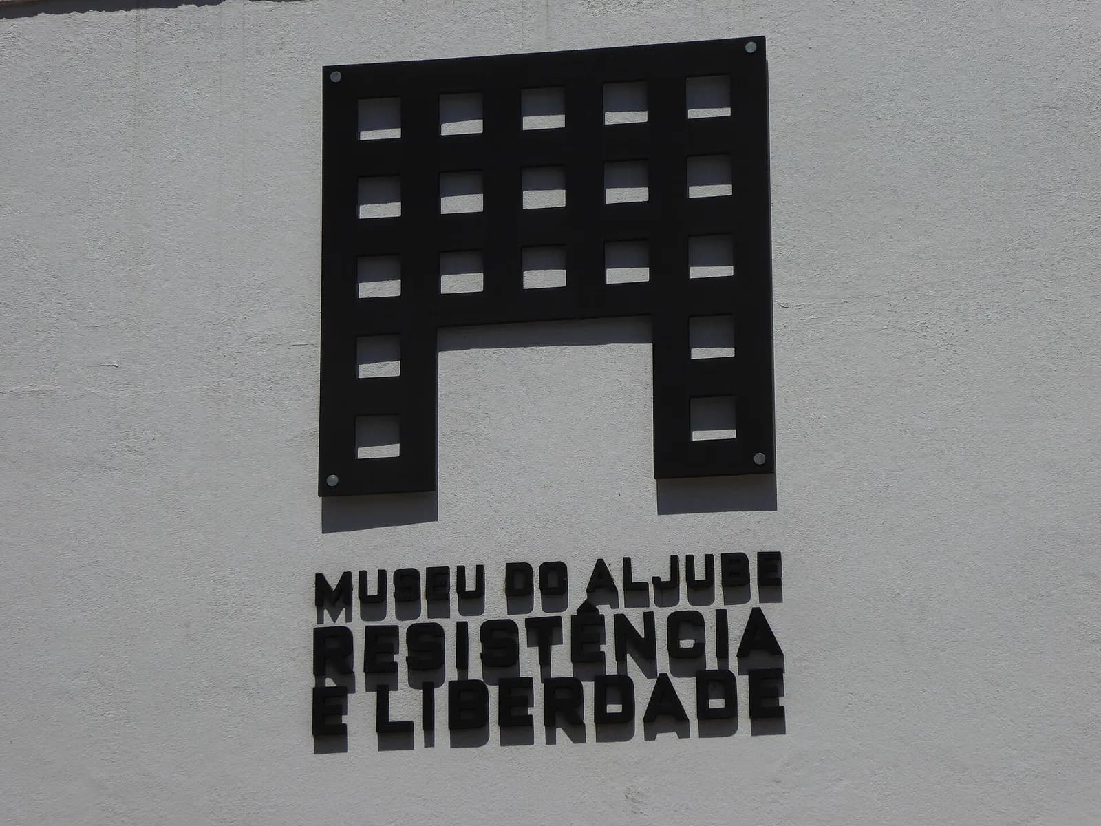 A black metal sign on a white wall: a grid of small square windows shaped like an arch over the words Museu do Aljube, Resistência e Liberdade in bold capitals.