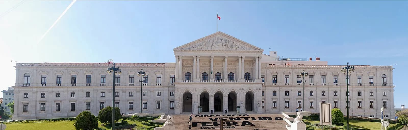Wide view of a long white neoclassical palace with a central columned portico and triangular pediment, a flag flying above it, and a broad flight of steps in front flanked by stone lions and lamp posts. Large black letters laid on the steps spell Arquitecto Ventura Terra.