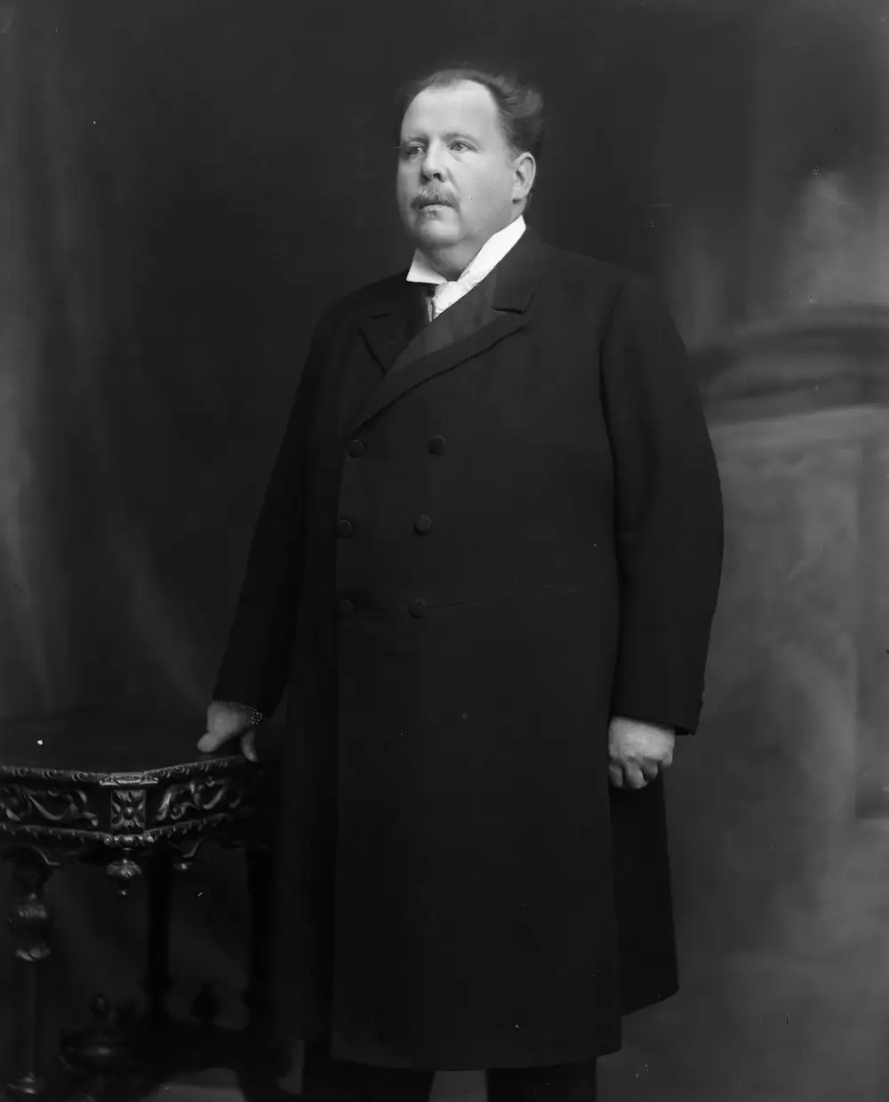 Studio portrait of a stout, moustached man in a long double-breasted black coat and high collar, standing with his right hand resting on a carved table.