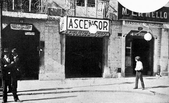 A shopfront doorway under a large sign reading Ascensor, with smaller lettering for the Biblioteca and Chiado. Two men in hats stand in the next doorway and a boy stands on the pavement.