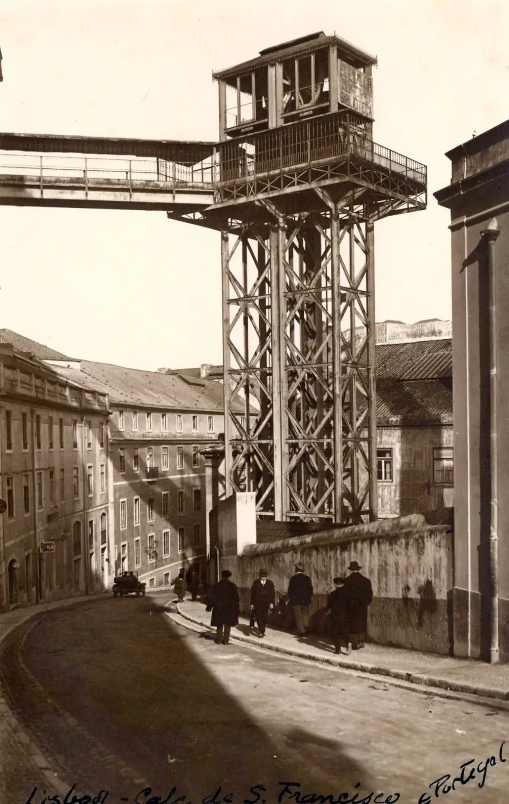 A tall open steel lattice tower topped by a glazed cabin house, joined at the top by a footbridge to the buildings on the left, rises above a sloping cobbled street where a few men in hats walk past.
