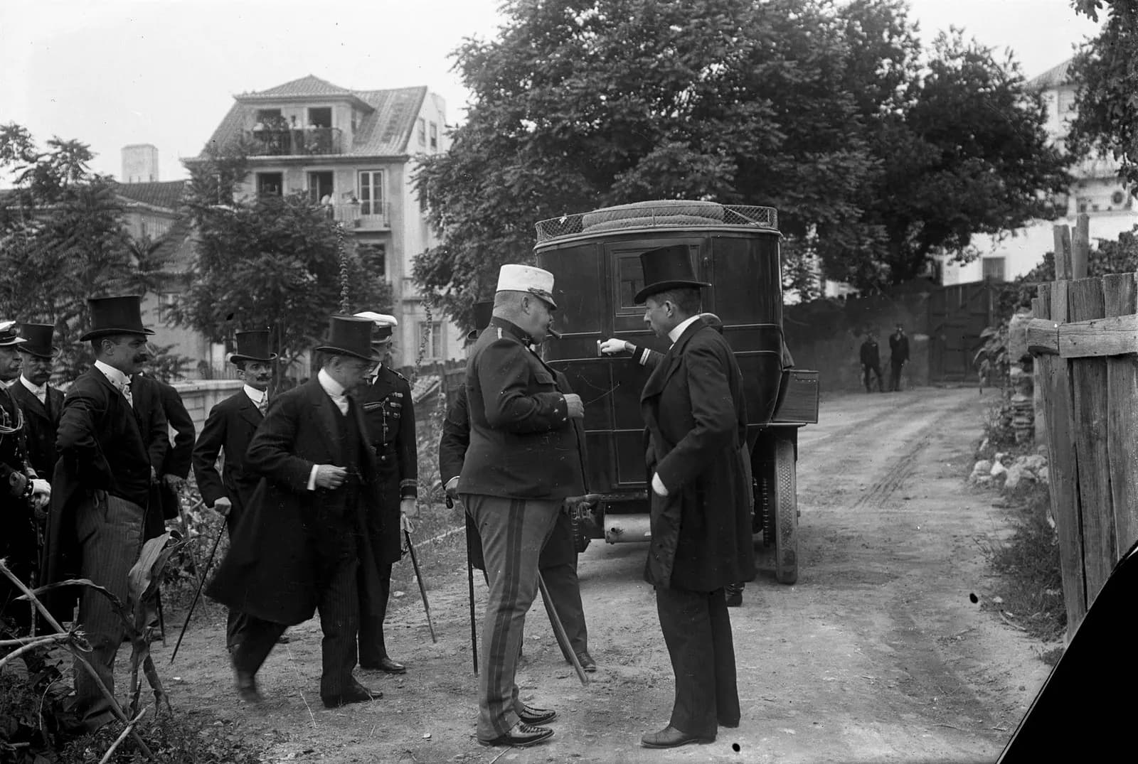 On a dirt road beside a boxy black motor car, a heavily built man in military uniform and a white-topped cap stands facing a man in a top hat and long dark coat. Several other men in top hats and frock coats stand to the left.