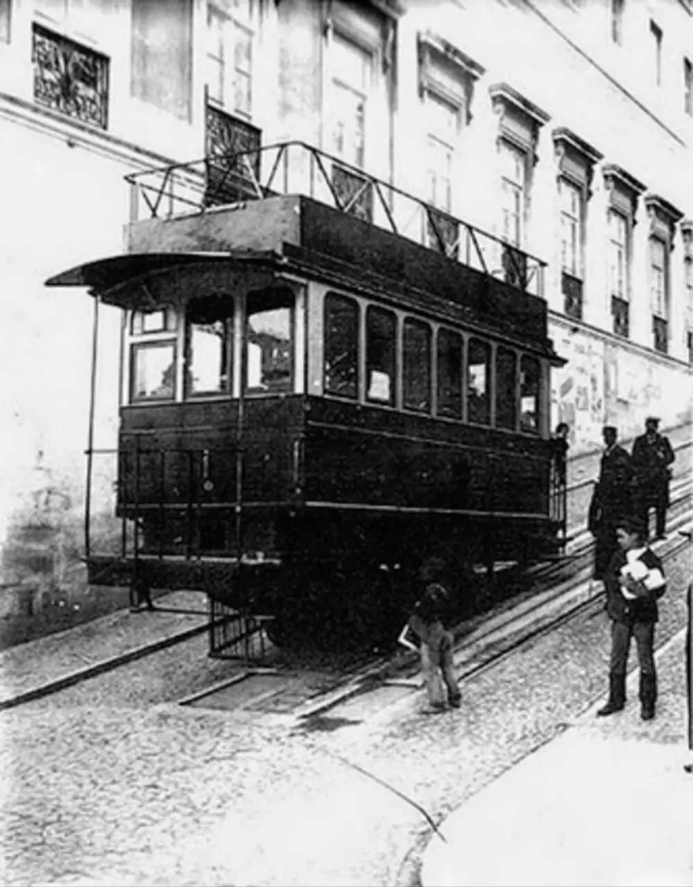 Black and white photograph of a dark two-storey tramcar with a railed upper deck and a curved canopy over its front platform, standing on a cobbled slope beside a tall building. Boys and men stand next to it, one boy holding a bundle of newspapers.