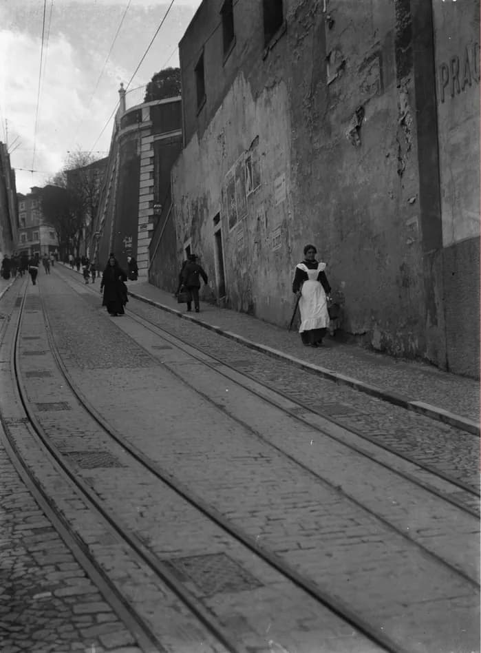 Black and white photograph looking up a steep cobbled street with two sets of rails set into the paving. Overhead wires run up the hill, a woman in a long white apron stands by a high stone wall and other pedestrians walk further up.