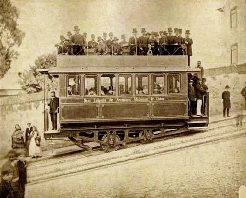 Sepia photograph of a long two-storey tramcar standing on a slope. Its side reads Nova Companhia dos Ascensores Mechanicos de Lisboa. Men in top hats pack the railed upper deck, more stand on the end platforms, and a toothed wheel is visible between the axles.