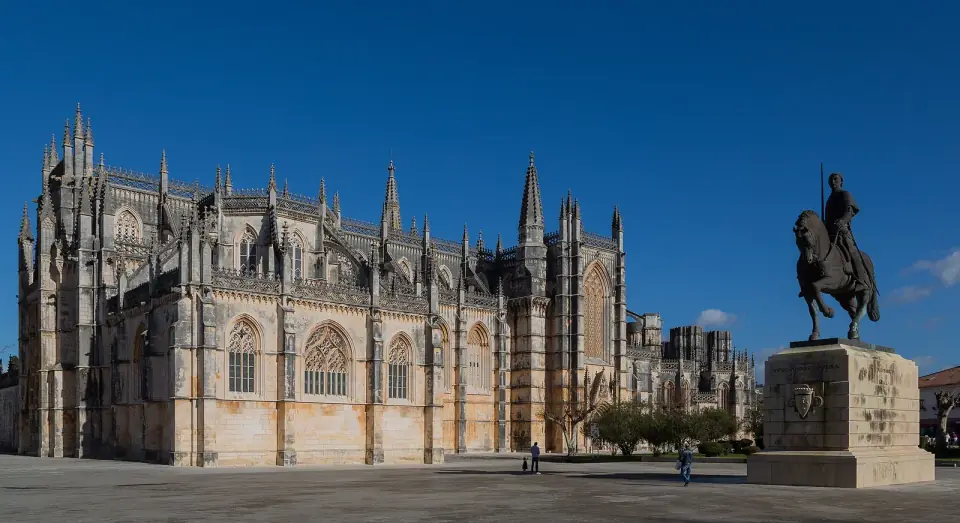 The long Gothic flank of a pale limestone monastery with pinnacles and tall windows, beside a bronze equestrian statue on a stone plinth, under a deep blue sky.