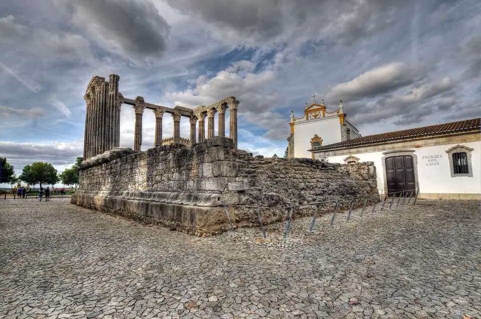 The granite podium and surviving Corinthian columns of a Roman temple on a cobbled square, with a whitewashed building beside it under a cloudy sky.