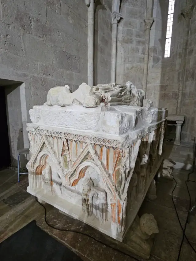 A large white stone tomb carved with pointed Gothic arches and traces of red paint, topped by a damaged reclining figure, inside a bare stone church.