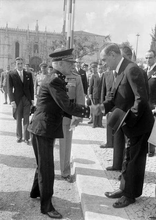 Black-and-white photograph of a man in military uniform and peaked cap shaking hands with a man in a dark suit holding his hat, with officials and a Gothic monastery behind them.