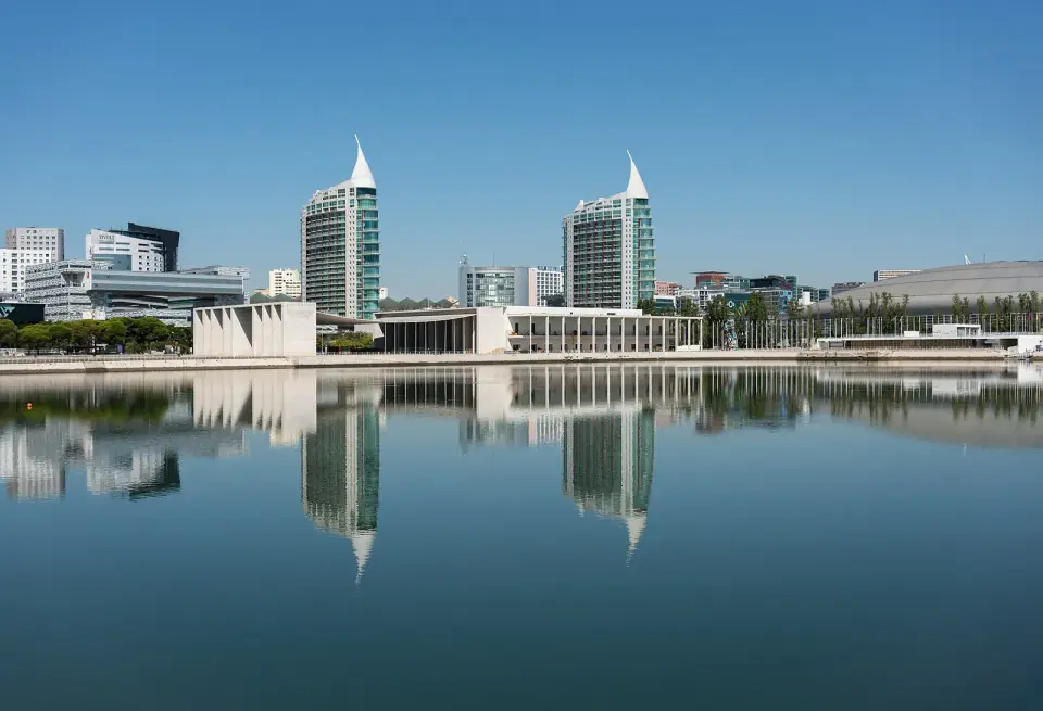 Two tall towers with pointed white tops and a row of low modern buildings reflected in the still water of a dock under a clear blue sky.