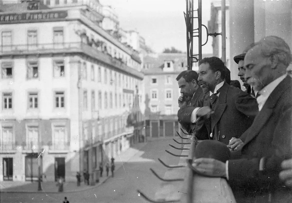 Black-and-white photograph of men in dark suits on a balcony, looking down on a street lined with tall buildings.
