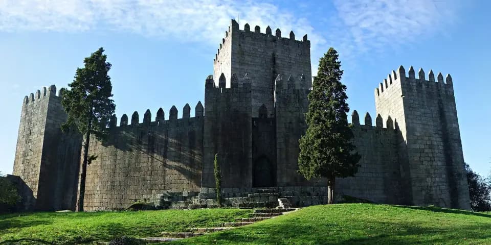 A square stone keep rising above crenellated castle walls and towers, with two dark cypress trees and a green lawn in front.