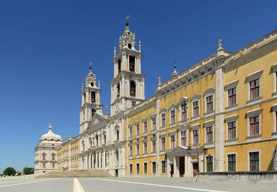 A long yellow and white Baroque palace front with rows of windows, and a church with two tall bell towers at its centre, under a clear blue sky.