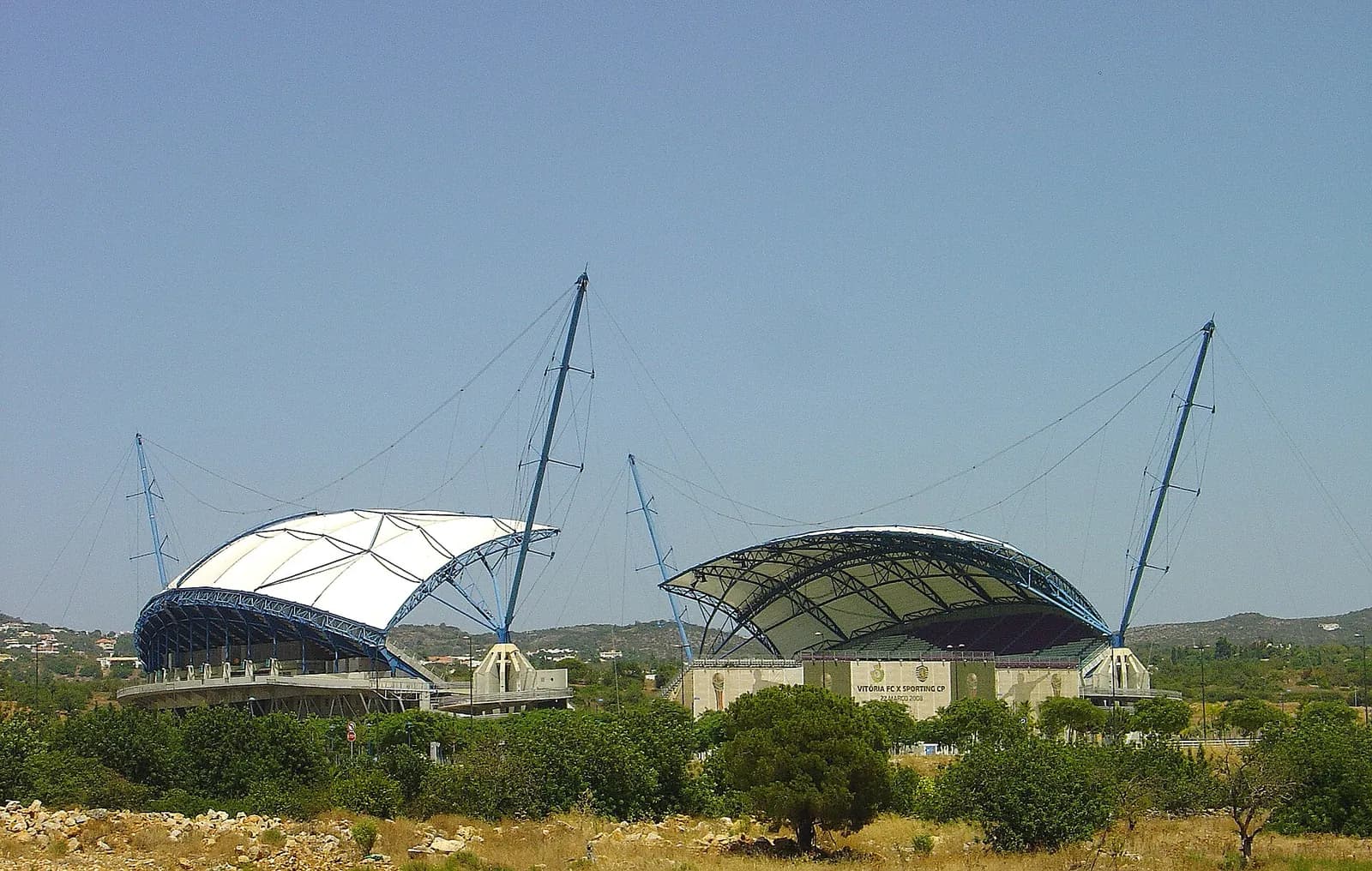 Two curved stand roofs, one white and one seen from beneath, each hung from tall blue masts by cables, rising behind scrub and pine trees under a clear sky.